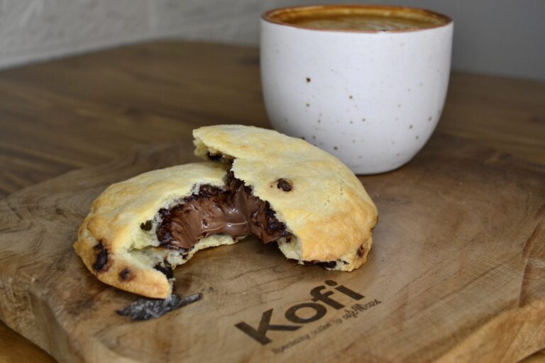 Un latte amb escuma i una cookie de xocolata sobre un plat de fusta amb el logo de Kofi gravat