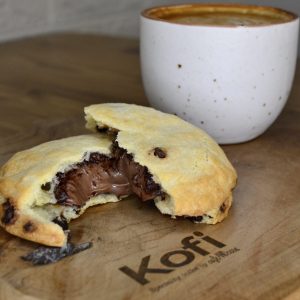 Un latte amb escuma i una cookie de xocolata sobre un plat de fusta amb el logo de Kofi gravat